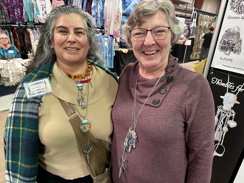 Two smiling women showcasing thimble accessories at a craft fair, surrounded by colorful fabric displays.